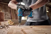 Person wearing safety equipment using a cordless drill to bore a hole in a wooden board.