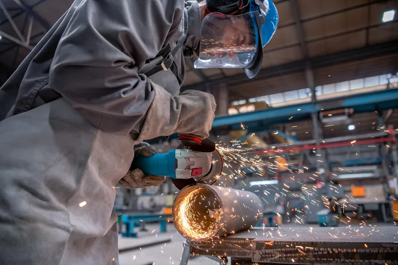 A person wearing safety equipment grinds a metal pipe with an angle grinder, sparks flying.