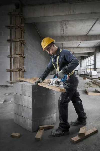 A person wearing safety equipment cuts wood with a circular saw on concrete blocks.