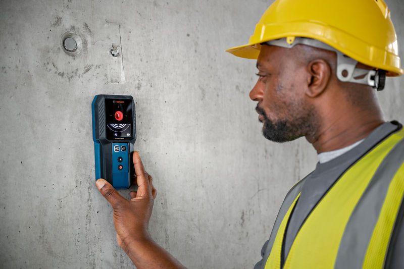 A person wearing safety equipment uses a detector to scan a concrete wall.