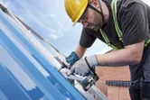 A person wearing safety equipment drives a screw into a blue metal roof with a power tool.