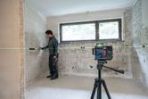 A person marks tiles on a wall using a laser leveling tool on a tripod.