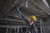 A person wearing safety equipment uses a reciprocating saw to cut a metal pipe overhead.