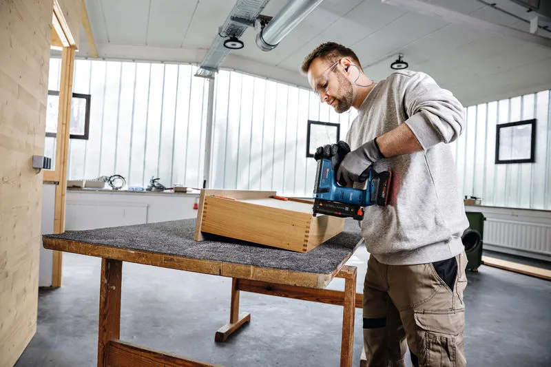 Person wearing safety equipment uses a cordless nail gun on a wooden box in a workshop.