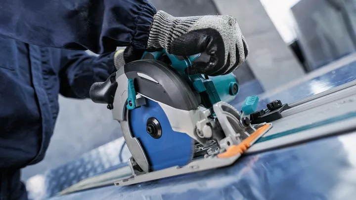 Person wearing safety equipment operates a circular saw to cut a sheet of material.