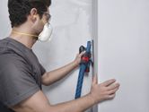 A person wearing safety equipment sands a wall with an orbital sander connected to a vacuum hose.