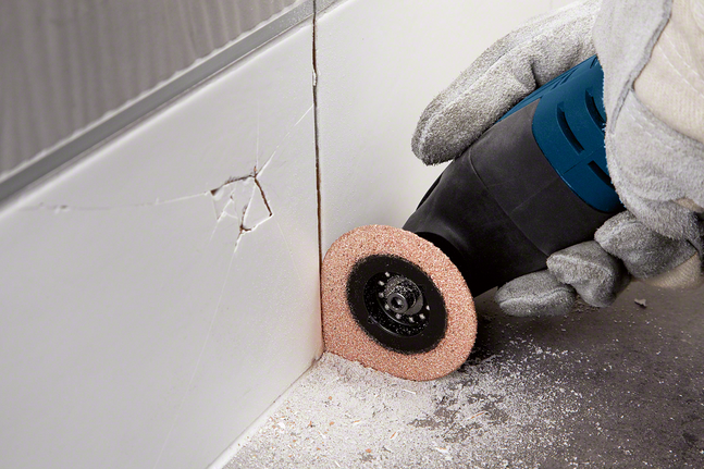 Person wearing safety equipment uses a power tool to cut into grout between wall tiles.