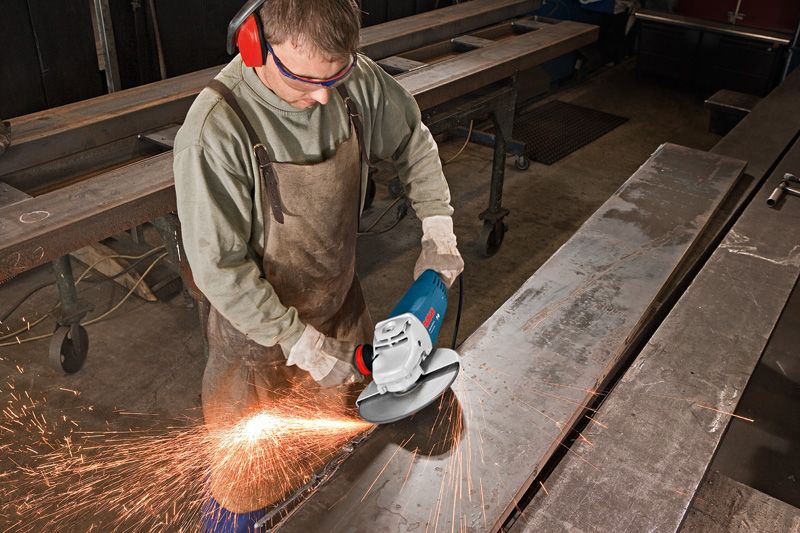 A person wearing safety equipment grinds a large metal beam, producing flying sparks.