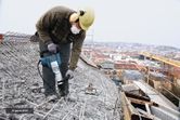 A person wearing safety equipment uses a demolition hammer to break concrete on a rooftop.