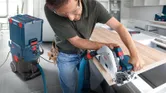A person wearing safety equipment cuts wood with a circular saw connected to a dust extractor.