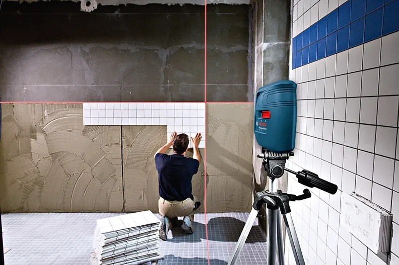 A person aligns wall tiles using a laser leveling tool on a tripod.
