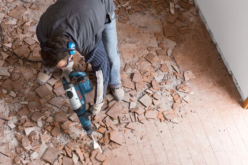 A person wearing safety equipment breaks floor tiles using a demolition hammer.