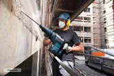 A person wearing safety equipment uses a demolition hammer to break concrete on a building site.