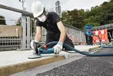 A person wearing safety equipment grinds concrete on a sidewalk with a connected grinder.