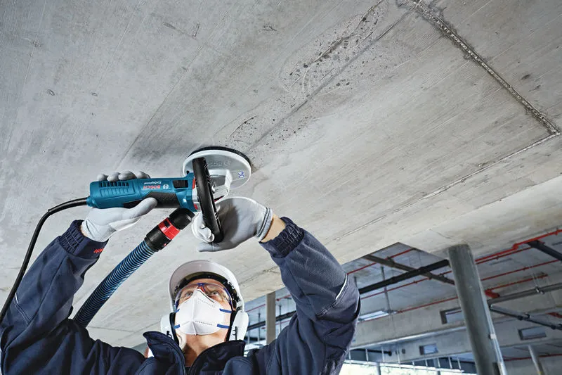 A person wearing safety equipment grinds a concrete ceiling with a concrete grinder.