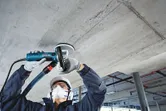 A person wearing safety equipment grinds a concrete ceiling with a concrete grinder.