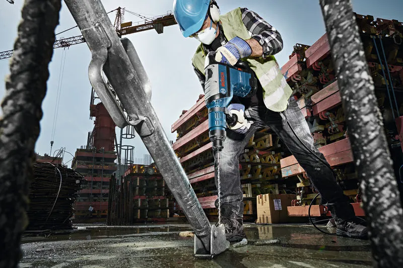 A worker wearing safety equipment uses a rotary hammer to drill into a concrete base at a construction site.