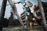 A worker wearing safety equipment uses a rotary hammer to drill into a concrete base at a construction site.