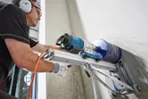 A person wearing safety equipment drills into a wall using a mounted drill stand.