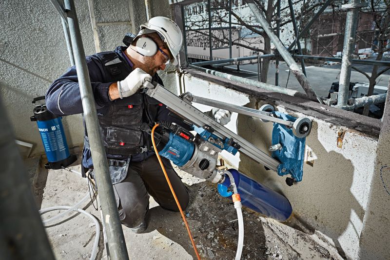 A person wearing safety equipment uses a drill stand to bore a hole in a concrete wall.