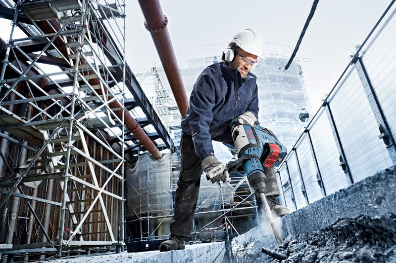 A person wearing safety equipment uses a cordless rotary hammer to drill concrete at a construction site.