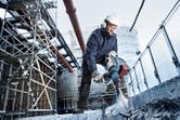 A person wearing safety equipment uses a cordless rotary hammer to drill concrete at a construction site.