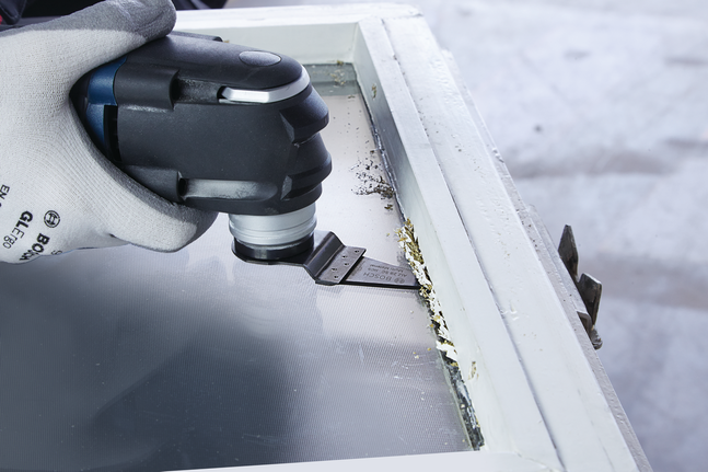 Person wearing safety equipment removes old paint from a window frame using a multi-tool.