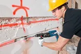 A person wearing safety equipment uses a rotary hammer to chisel a groove in a concrete wall.