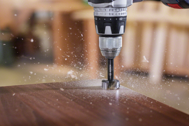Drill boring a hole in a wooden board while wood shavings scatter.