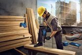 A person wearing safety equipment drills into a wooden beam at a construction site.