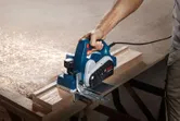 A person uses an electric planer to smooth a wooden board on a workbench.