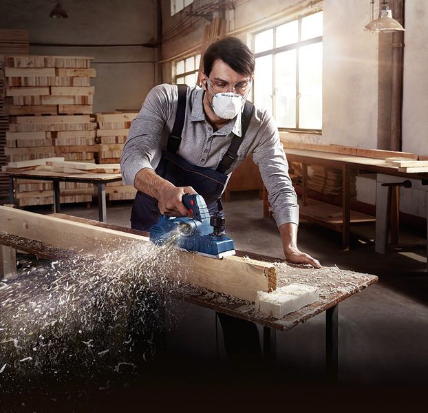 A person wearing safety equipment planes a wooden beam in a workshop.