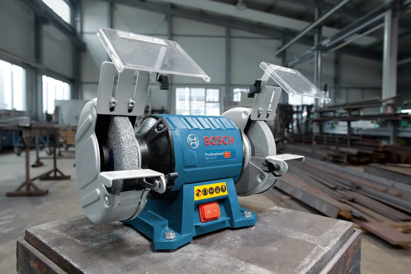 Double-wheeled bench grinder with protective shields on a workbench in an industrial workshop.