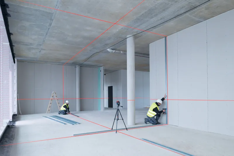 Two workers wearing safety equipment align drywall using a laser leveling tool.