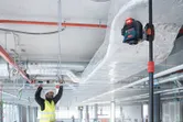 A person wearing safety equipment aligns ceiling structures using a laser leveling tool.