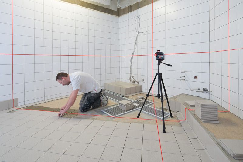 Person installing tiles in a bathroom, using a laser leveling tool on a tripod.