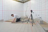 Person installing tiles in a bathroom, using a laser leveling tool on a tripod.