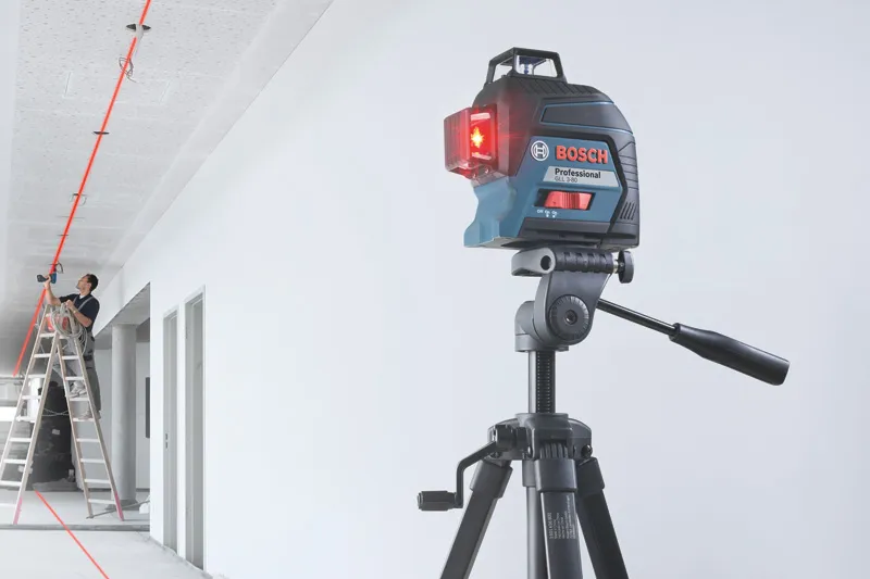 Laser leveling tool on a tripod projects red lines while a person works on a ceiling.