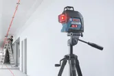 Laser leveling tool on a tripod projects red lines while a person works on a ceiling.