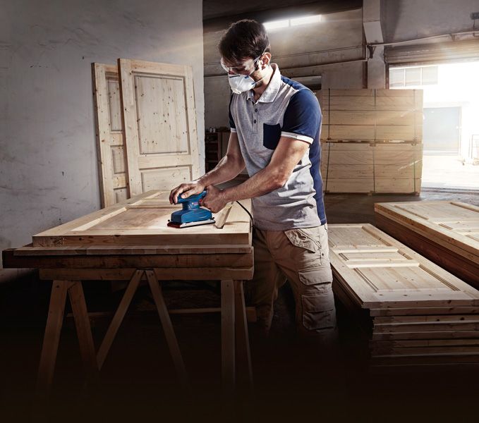 A person wearing safety equipment sands a wooden door with an orbital sander.