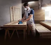 A person wearing safety equipment sands a wooden door with an orbital sander.