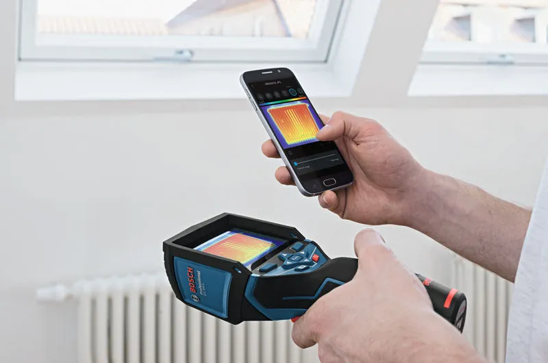 Person holds a thermo camera while viewing heat readout on a smartphone indoors.