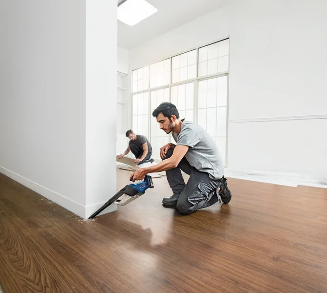 A person cuts flooring near a wall with a cordless saw while another installs floor panels.