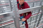 A person wearing safety equipment uses a power drill on a metal beam in a warehouse.