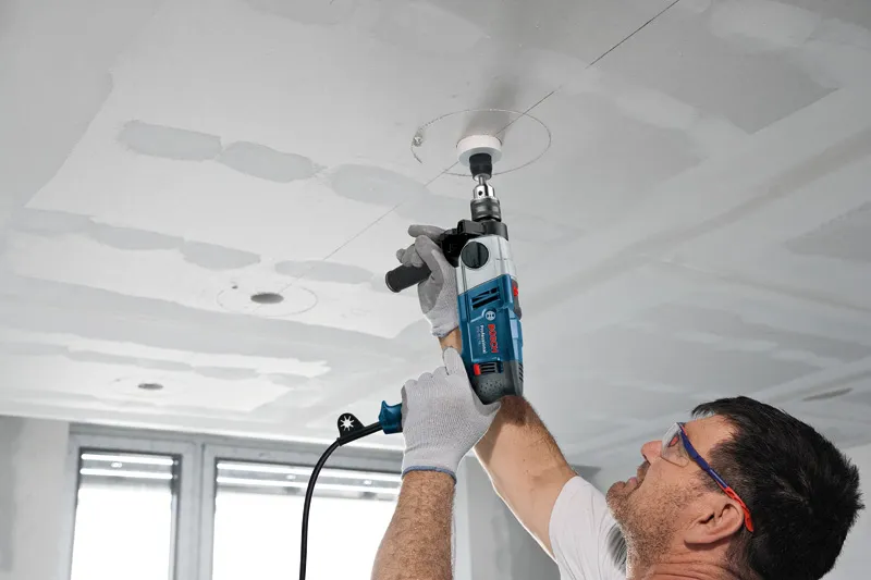 A person wearing safety equipment drills a circular hole into a ceiling with a power tool.