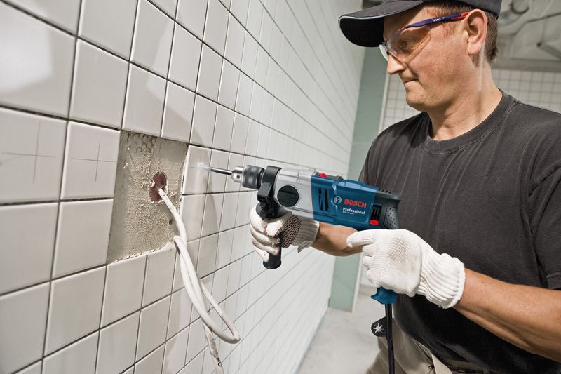 A person wearing safety equipment drills into tiled wall above exposed cables.