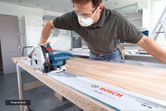 A person wearing safety equipment uses a plunge saw to cut wood along a guide rail.