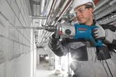 Electric impact drill with auxiliary handle and long masonry bit attached.