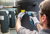 A person wearing safety equipment uses a heat gun to apply a label to a large tank.