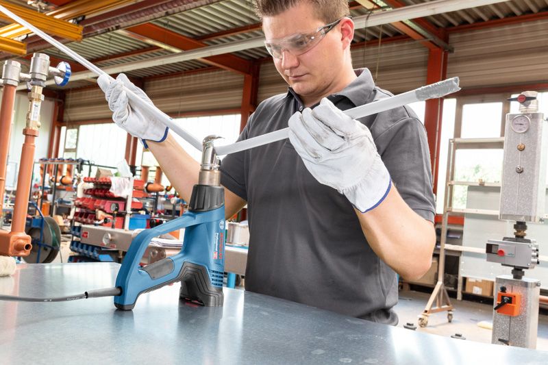 A person wearing safety equipment heats a plastic pipe with a heat gun in a workshop.